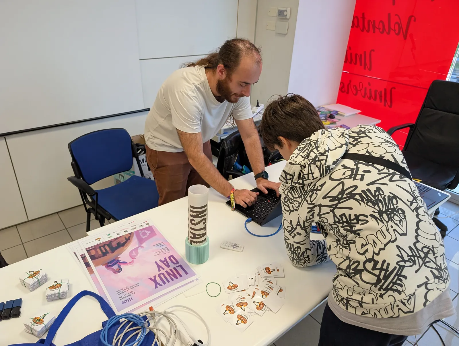 A volunteer and a teenager bent over the same laptop, on a table with the Linux Day poster, stickers and network cables