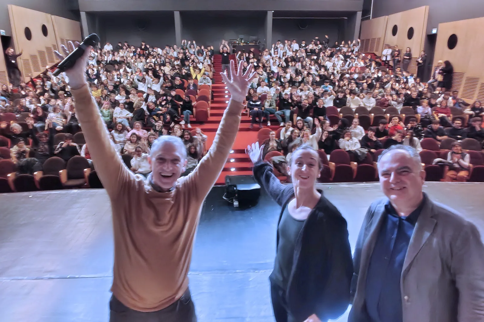 A hall full of young people and adults waving with their arms raised; in the foreground, on stage, three smiling organisers