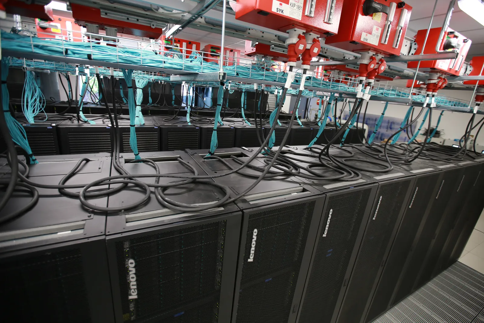 Rows of black supercomputer cabinets with bundles of light blue cables above them, in a machine room