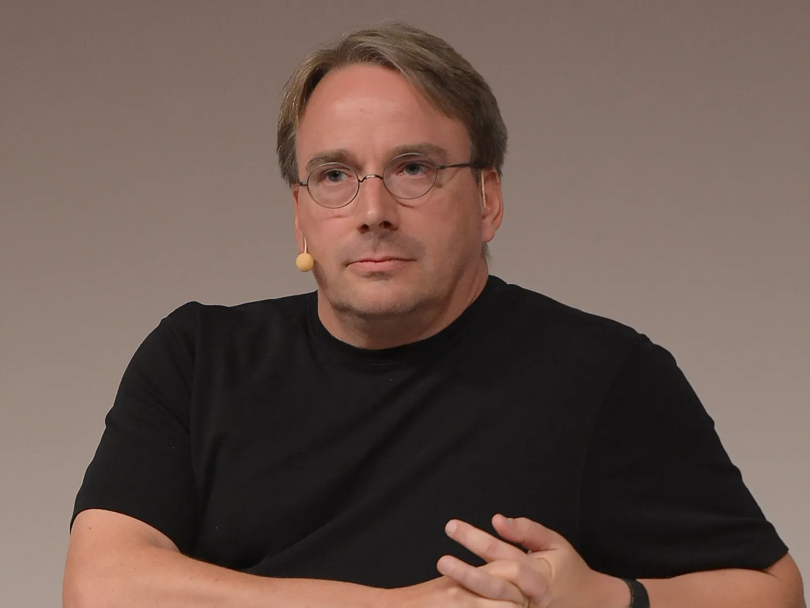 Linus Torvalds sitting on a small white sofa on stage at LinuxCon Europe 2014, in a black T-shirt and round glasses